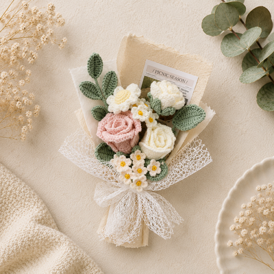 A crocheted flower bouquet with pink and white roses, white daisies, and green leafy accents, wrapped in cream paper and lace ribbon on a beige textured background with dried flowers and eucalyptus leaves.
