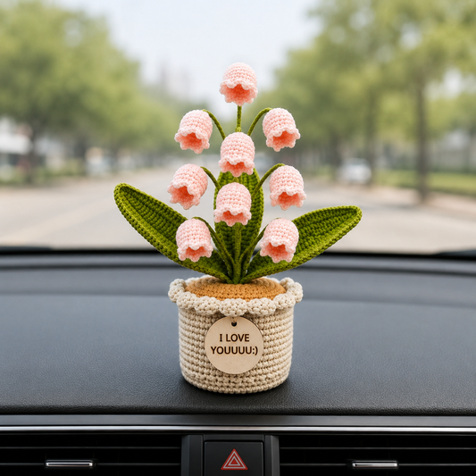 A crocheted bell-shaped flower arrangement in a small beige pot with the text 'I LOVE YOUUUU!' on the front, displayed on a car dashboard with a blurred outdoor street scene in the background.