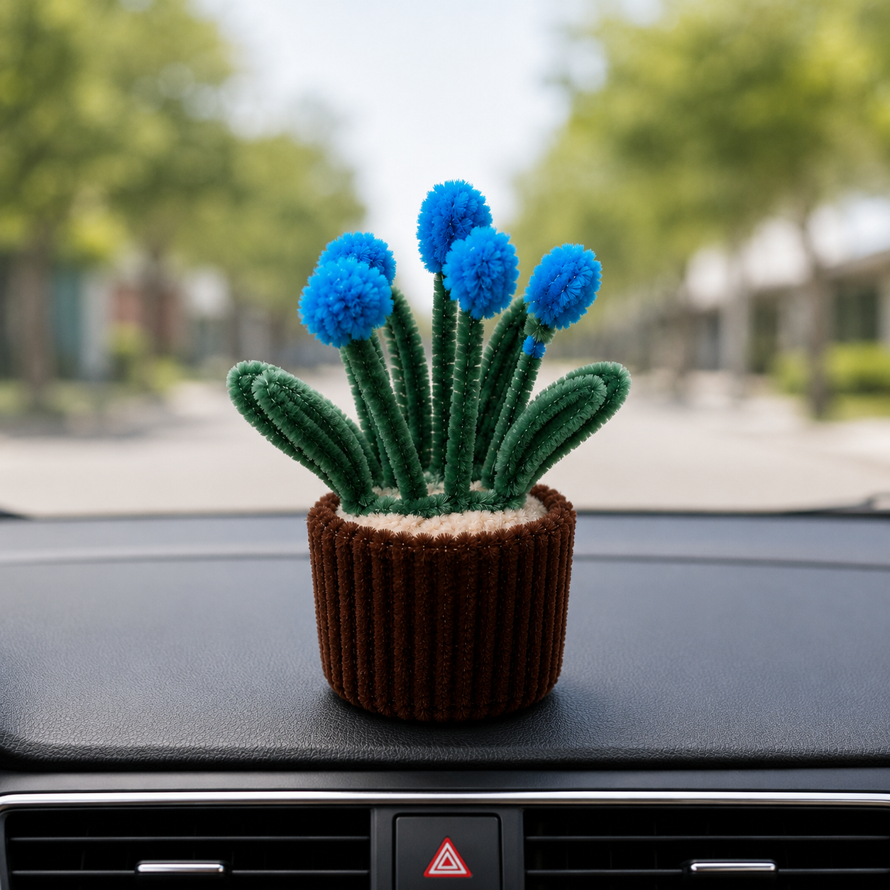 A small cactus-shaped decorative item with blue pom-pom flowers and green textured stems, placed on a car dashboard with a blurred outdoor street background.