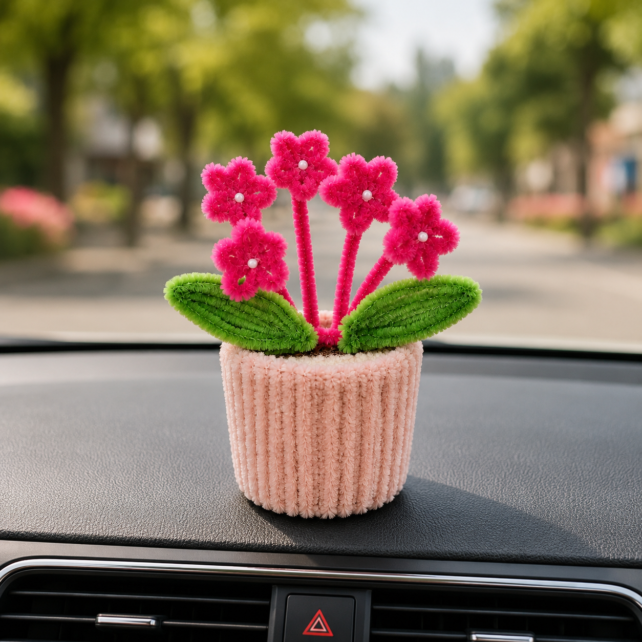 A small decorative flower arrangement in a pink ribbed pot sits on a car dashboard with a blurred outdoor street scene behind it. The flowers are bright pink with green leaves and white centers.