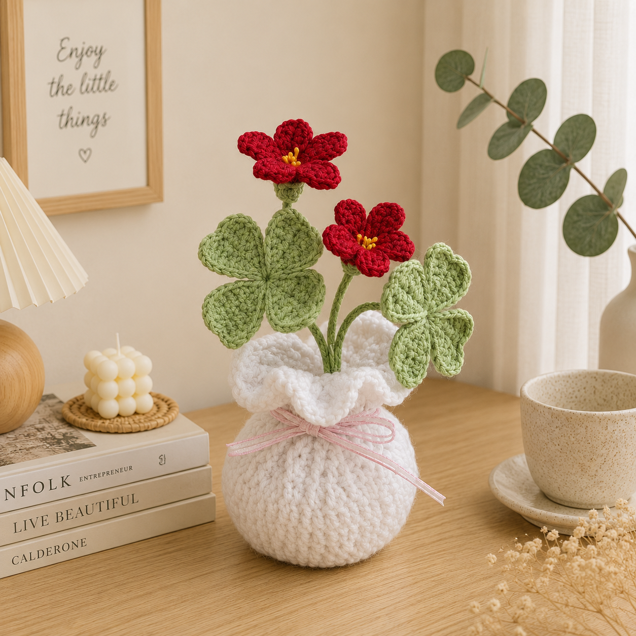 A crocheted white flower vase with a pink ribbon, holding two red crochet flowers with green four-leaf clover-style leaves, styled on a wooden table with books, a candle, a cup, and framed wall art.