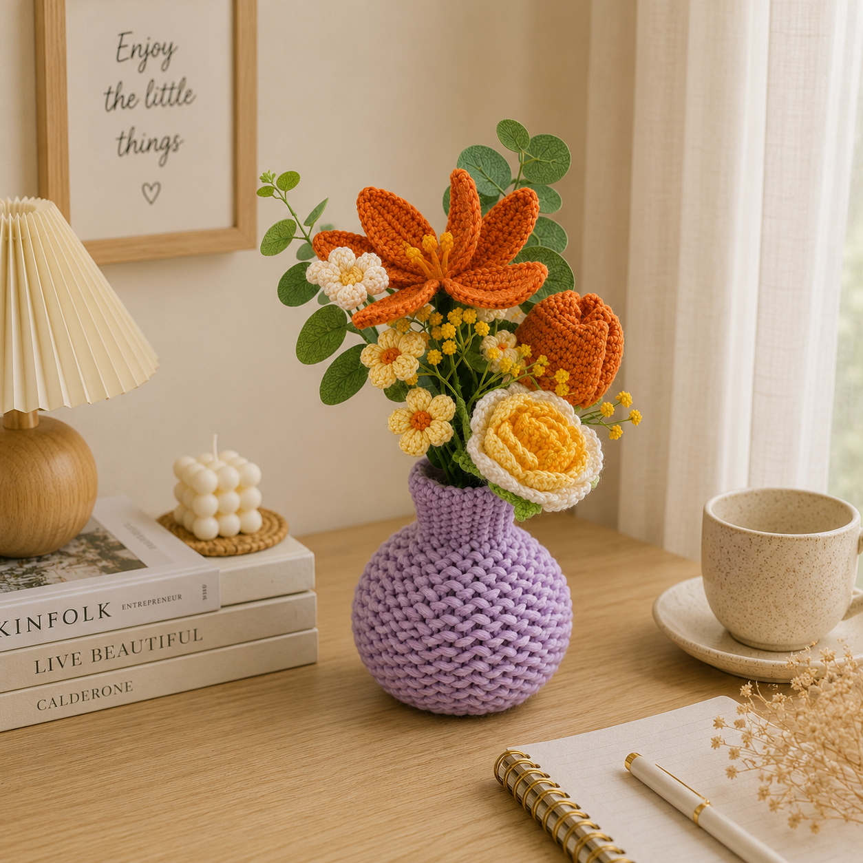 A crocheted lavender vase filled with a colorful handmade flower arrangement, styled on a wooden table in a warm, cozy room setting with a lamp, books, and a cup.
