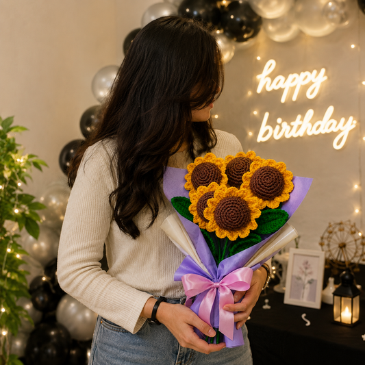A woman in a cream ribbed sweater and blue jeans holds a bouquet of crocheted sunflowers wrapped in purple and white paper with a purple bow. The background features a warm 'happy birthday' neon sign, black and silver balloon garlands, and soft string lights.
