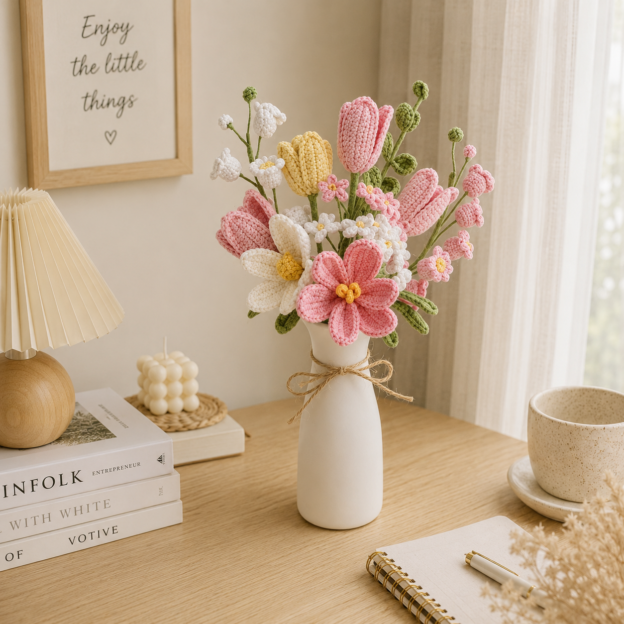 Handmade crocheted artificial flowers in pink, white, and yellow arranged in a small white ceramic vase on a wooden table, with a lamp, books, and a notebook nearby.