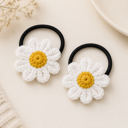 Two black elastic hair ties decorated with crocheted daisy flowers in white and yellow, arranged on a light beige surface with small dried floral props.