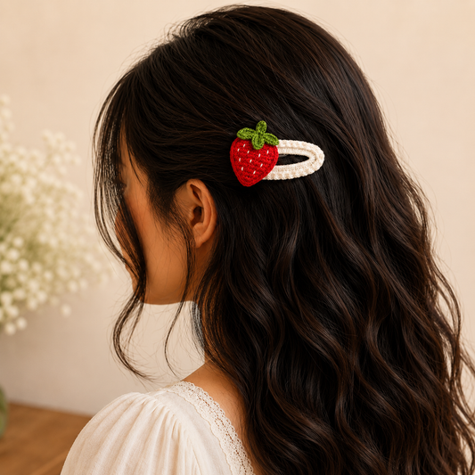 A strawberry-shaped hair clip with a white pearl-like band and a red strawberry charm with green leaves, shown clipped to the side of a woman's dark wavy hair.