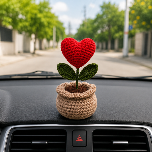 A small crocheted flower ornament with a red heart-shaped bloom, green leaves, and a beige pot sits on a car dashboard with a blurred street scene in the background.