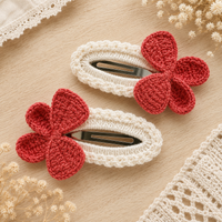 Two crocheted hair clips with white and red flower designs, displayed on a light wood surface with decorative lace and dried flowers.