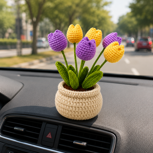 A crocheted tulip flower arrangement in a small beige pot, placed on a car dashboard with a blurred street scene visible through the windshield.