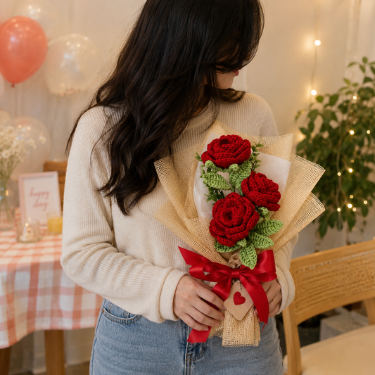A person in a cream ribbed sweater holds a bouquet of three red crochet roses wrapped in beige paper with a red ribbon and heart tag, set in a cozy indoor room with balloons and string lights.