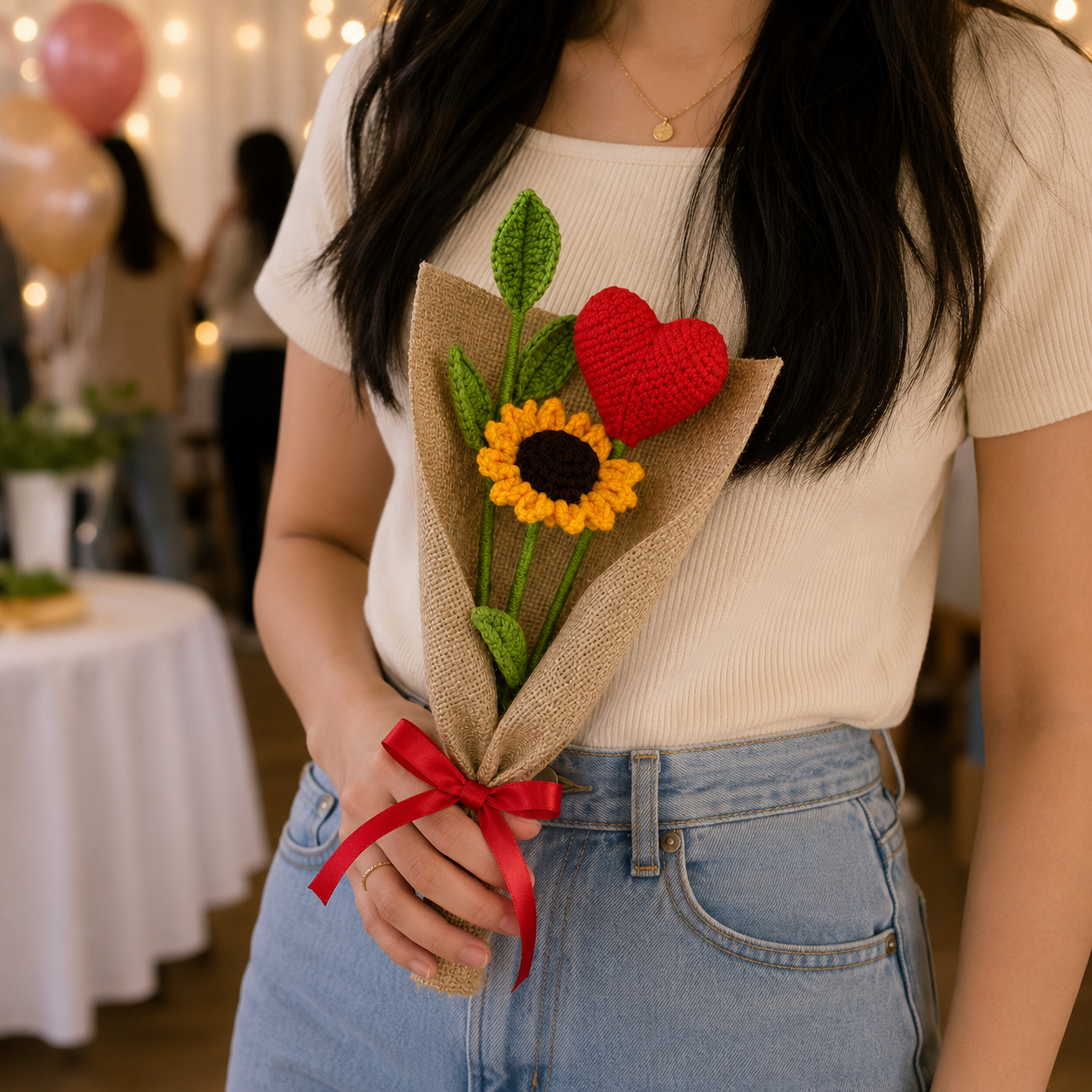 A person in a cream ribbed top and light blue jeans holds a small handmade crochet bouquet with a yellow sunflower, green leaves, a red heart, and burlap wrapping in a softly lit indoor setting.