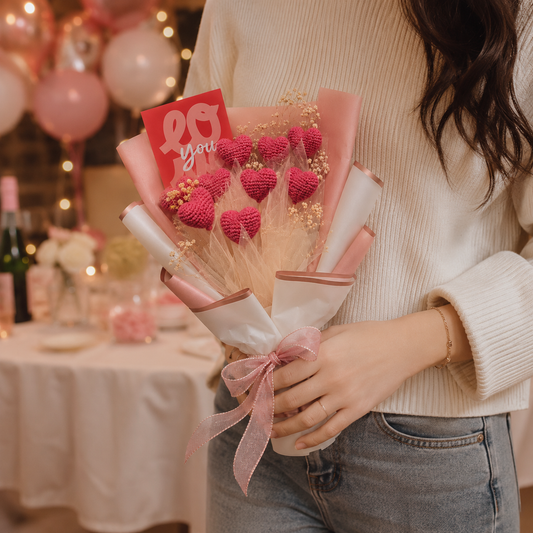 A person in a cream ribbed sweater holds a small bouquet made of pink crocheted hearts with a red card that says 'love you' in a softly lit indoor setting with balloons and table decorations in the background.