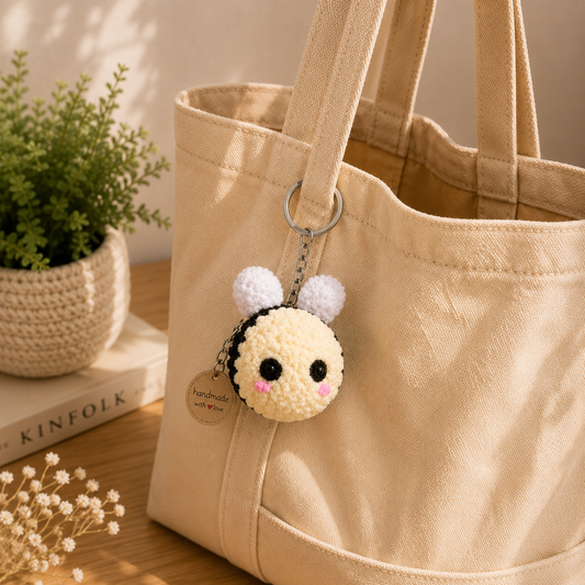 A beige canvas tote bag on a wooden surface with a small crocheted bee keychain hanging from a metal ring. The tote bag is styled with a potted plant and a book in a warm, natural setting.