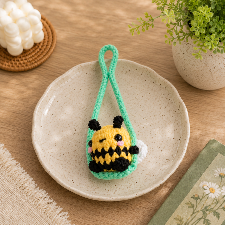 A small crocheted bumble bee keychain or bag charm sits on a speckled ceramic plate on a wooden table. The charm is yellow and black with pink cheeks and white wings, hanging from a mint green loop.