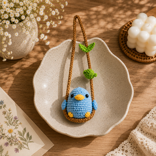 A crocheted blue bird hanging from a brown swing with green leaves, displayed on a beige ceramic dish over a wooden surface with floral props.