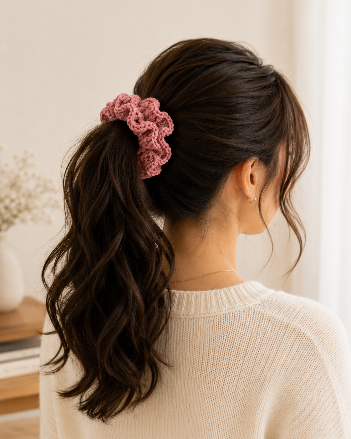 A woman shown from the back wearing a dusty pink crocheted scrunchie ponytail in her dark brown hair, wearing a cream knit sweater in a brightly lit indoor setting.