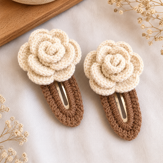 Two crocheted hair clips styled as cream-colored flowers on brown bands, displayed on a textured white fabric with dried floral accents.