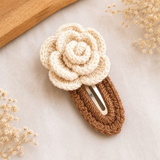 A crocheted hair claw clip styled with a cream-colored flower on top and a brown textured base, resting on a light beige linen fabric with dried flowers and a wooden board in the background.