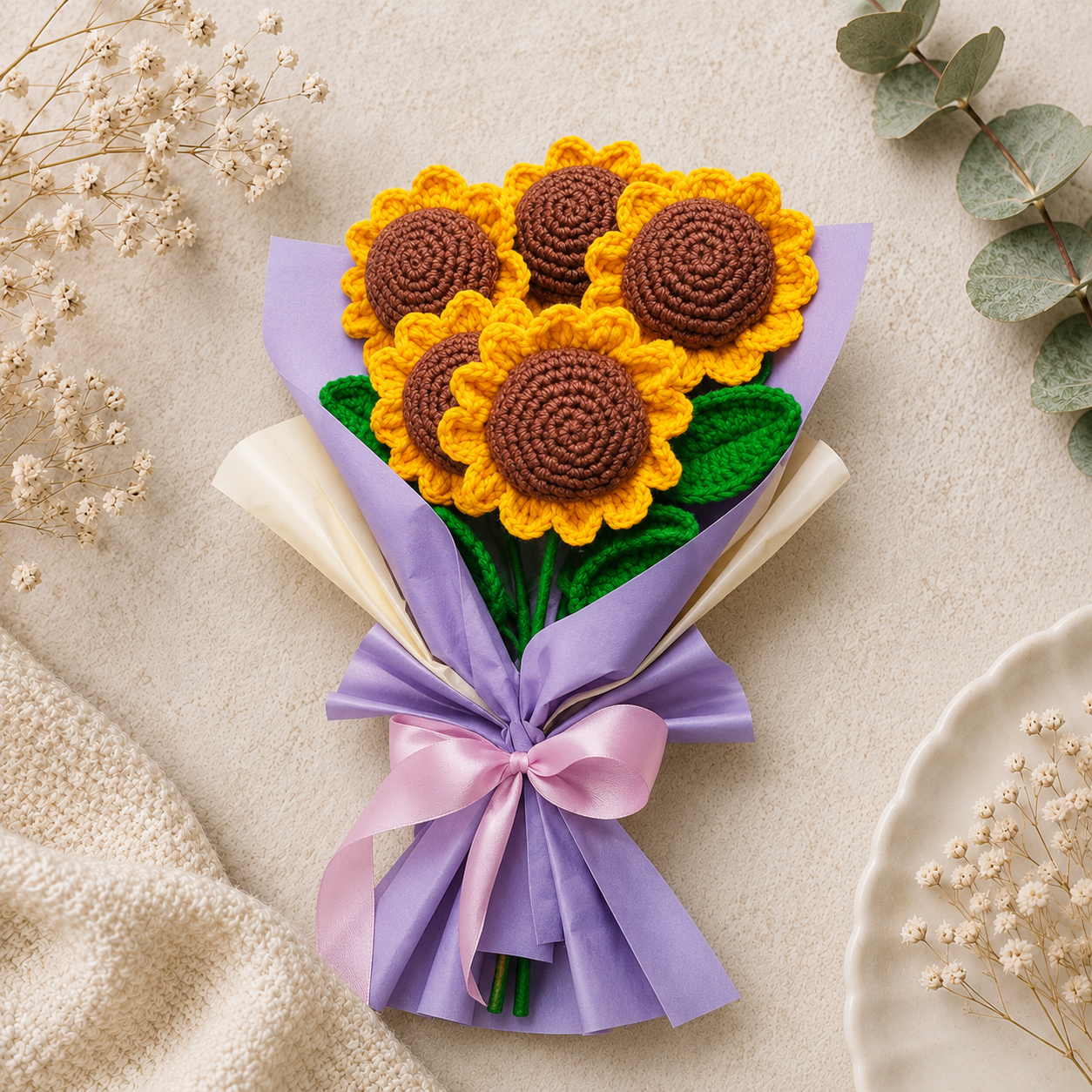 A handmade crochet sunflower bouquet wrapped in lavender and cream paper with a pink ribbon, styled on a neutral textured background with dried flowers and greenery.