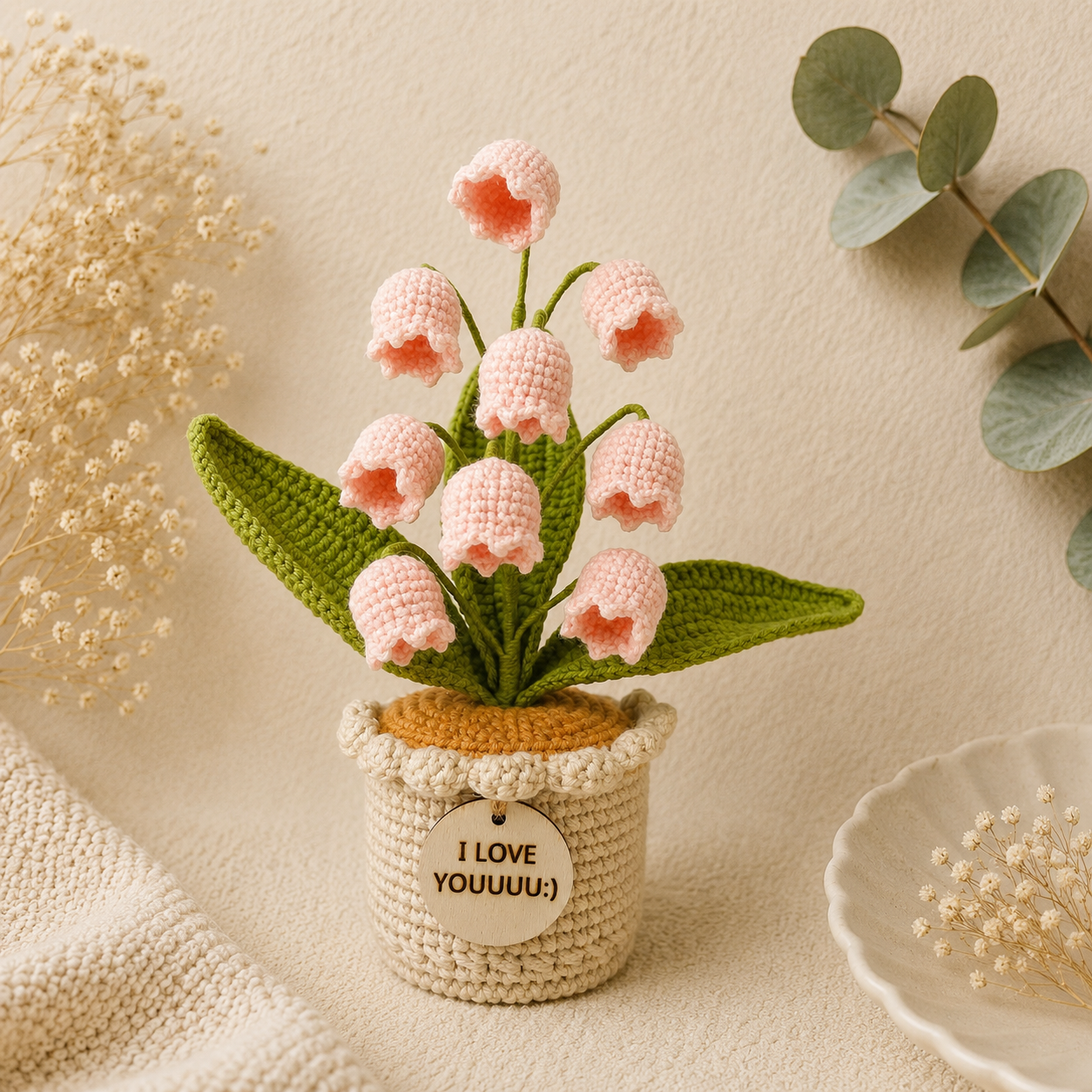 A crocheted floral arrangement featuring pink bell-shaped flowers with green leaves in a beige knitted pot, styled on a cream fabric surface with dried flowers and greenery in the background.