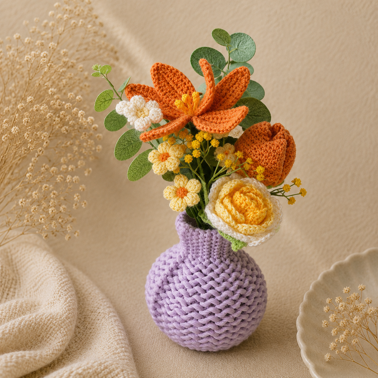 A crocheted lavender vase cover holding a handmade floral arrangement with orange lilies, yellow and white roses, and green leaves, styled on a beige fabric surface with dried flowers and a ceramic dish.