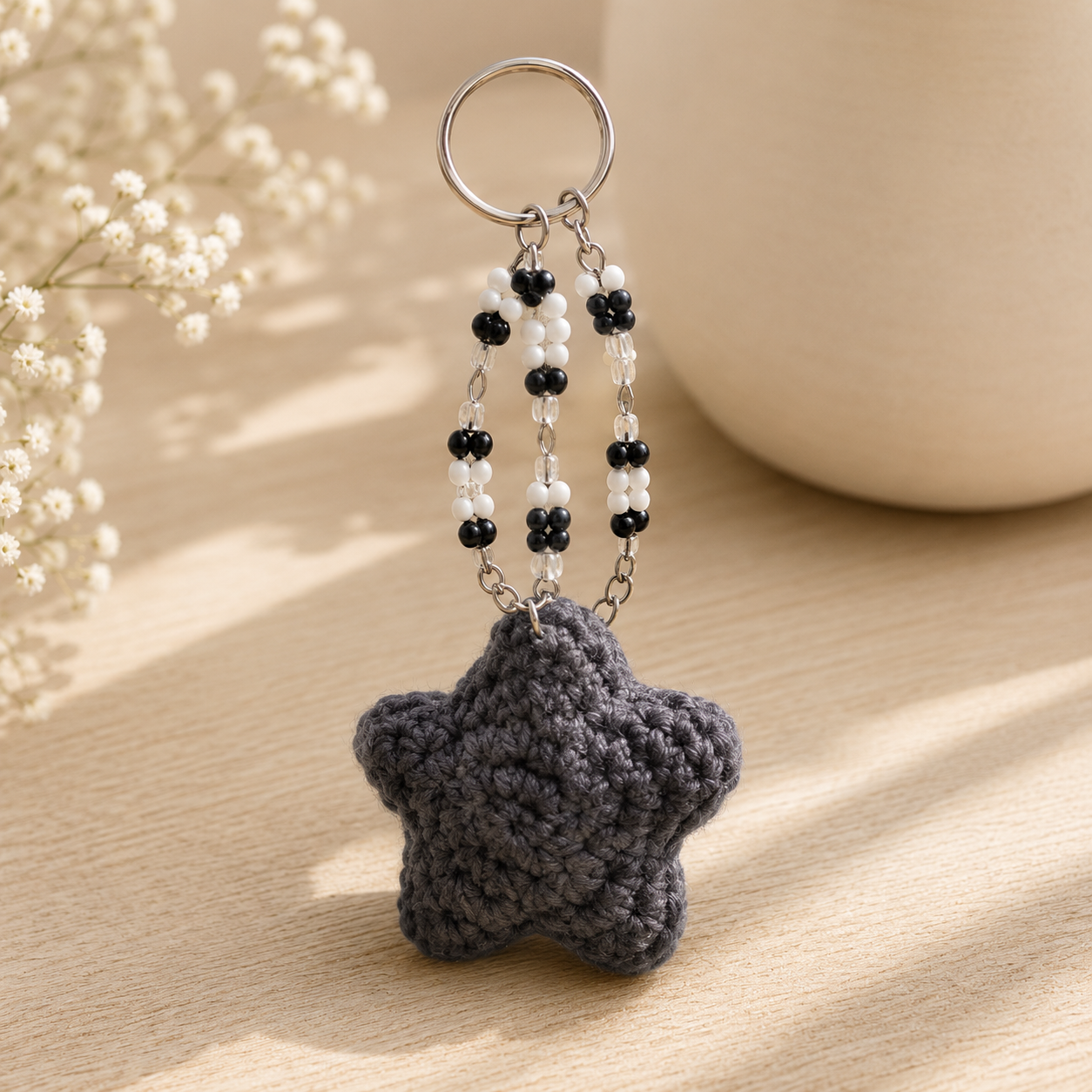 A dark gray crocheted star-shaped keychain with black and white beaded chains and a silver metal key ring, displayed on a light wooden surface with soft-focus background elements.