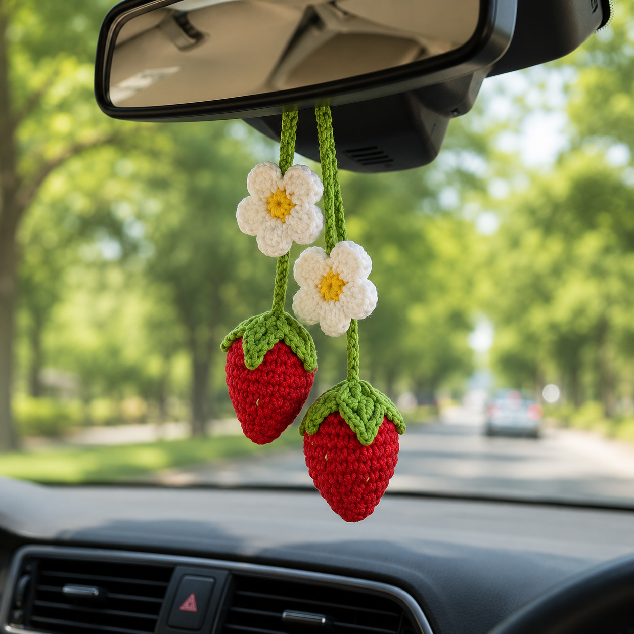 A pair of crocheted strawberry car air fresheners hanging from the rearview mirror inside a car, with a blurred green tree-lined road in the background.