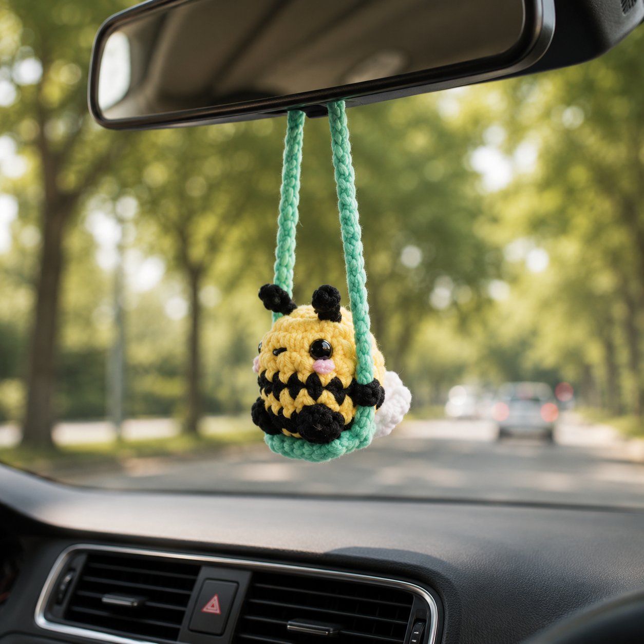 A small crocheted bee-shaped car air freshener hangs from a green braided strap attached to a rear-view mirror inside a car, with a blurred tree-lined road in the background.
