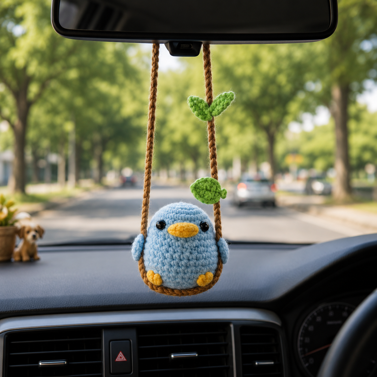 A crocheted blue bird car hanging ornament with yellow beak and feet, suspended from a brown rope swing with green leaves, displayed on a car dashboard with a blurred tree-lined road in the background.