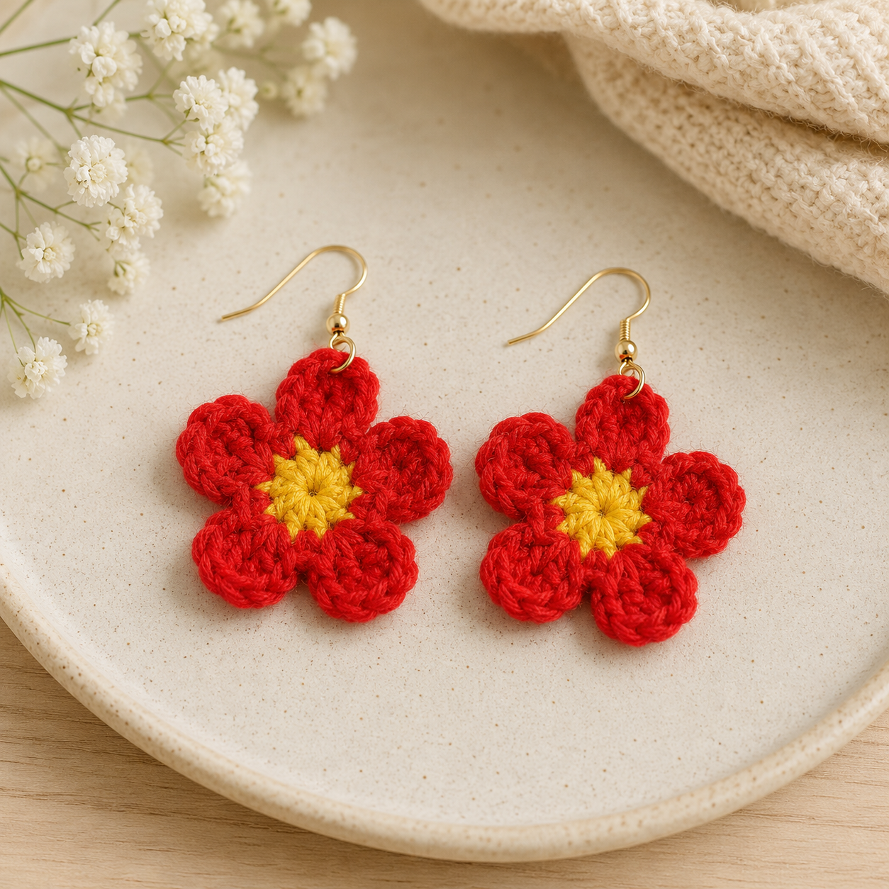 A pair of handmade red crochet flower drop earrings with yellow centers and gold-tone hooks, displayed on a light beige ceramic plate with small white flowers and a cream knit fabric in the background.