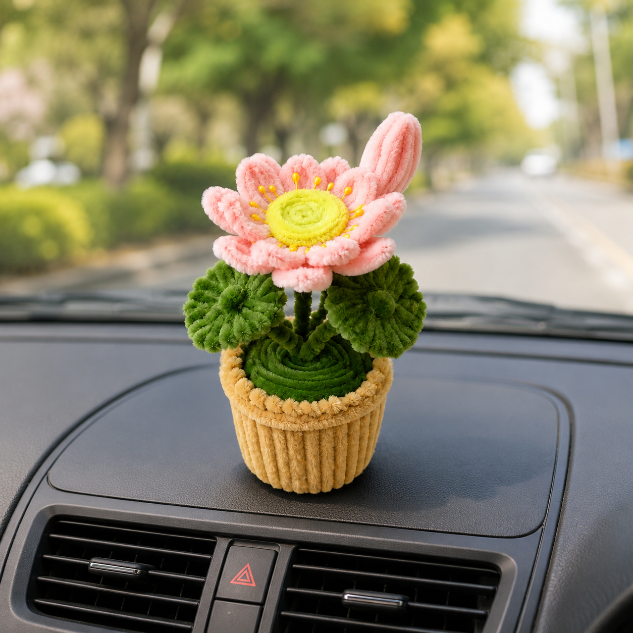 A small crocheted flower ornament in a ribbed pot sits on a car dashboard with a blurred road and trees in the background.