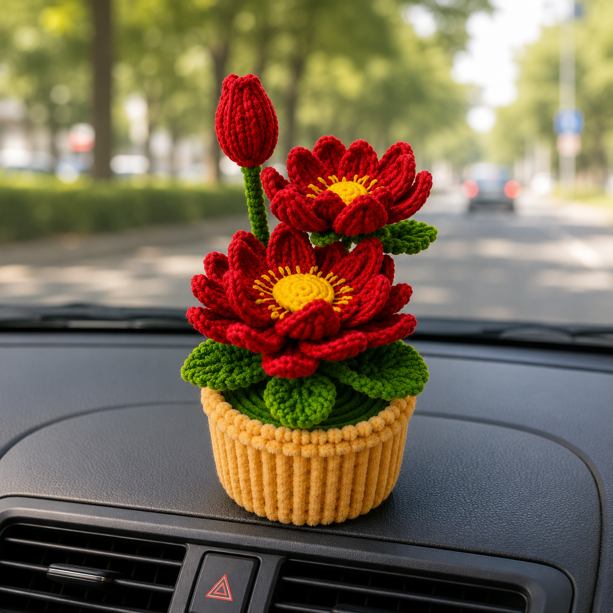 A crocheted flower pot ornament with red flowers, green leaves, and a yellow pot, placed on a car dashboard with a blurred street background.