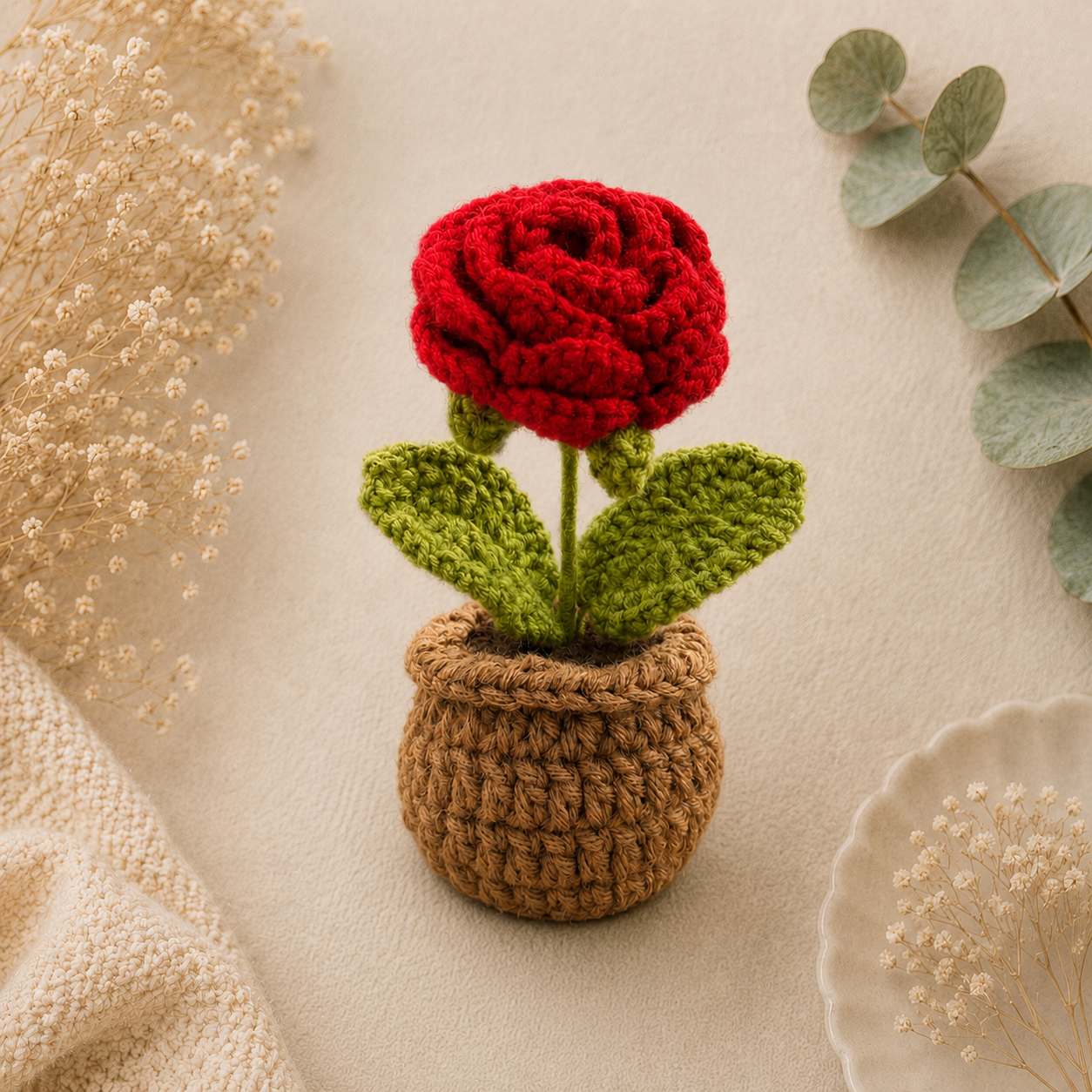 A crocheted artificial flower with a red rose bloom, green leaves, and a brown textured pot, styled on a beige fabric background with dried flowers and eucalyptus leaves.