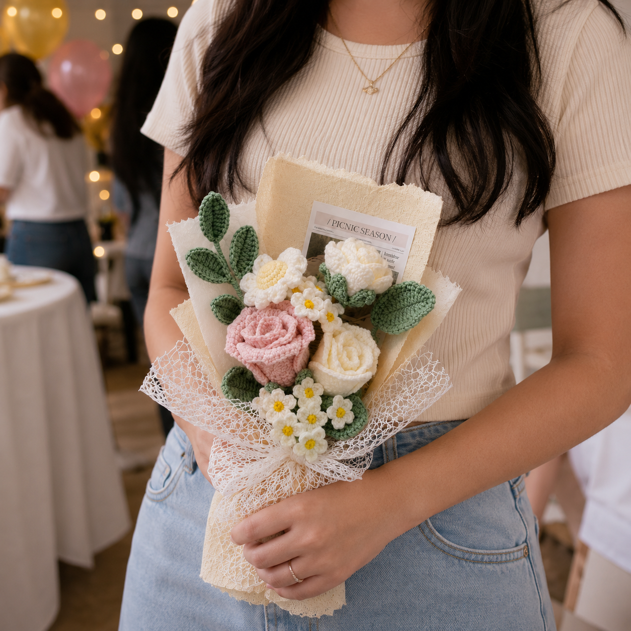 A person holding a handmade crochet flower bouquet with pink and white roses, white daisies, green leaves, and cream paper wrapping in a party setting.