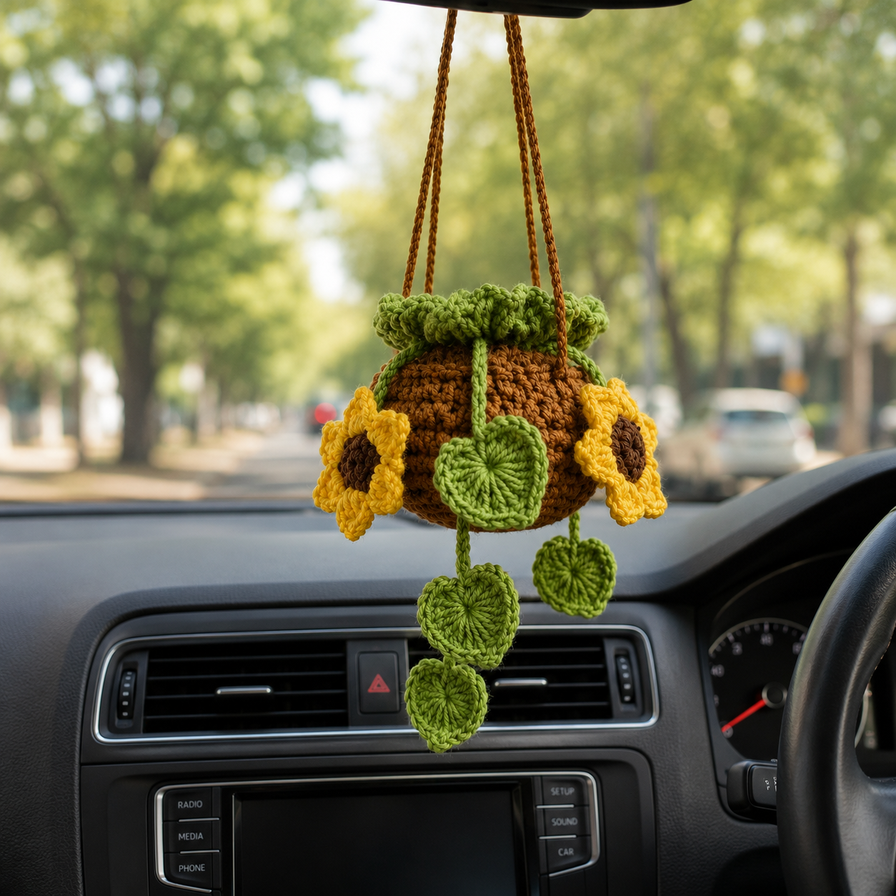 A crocheted sunflower car hanging ornament is displayed on a car dashboard with a blurred street background visible through the windshield.