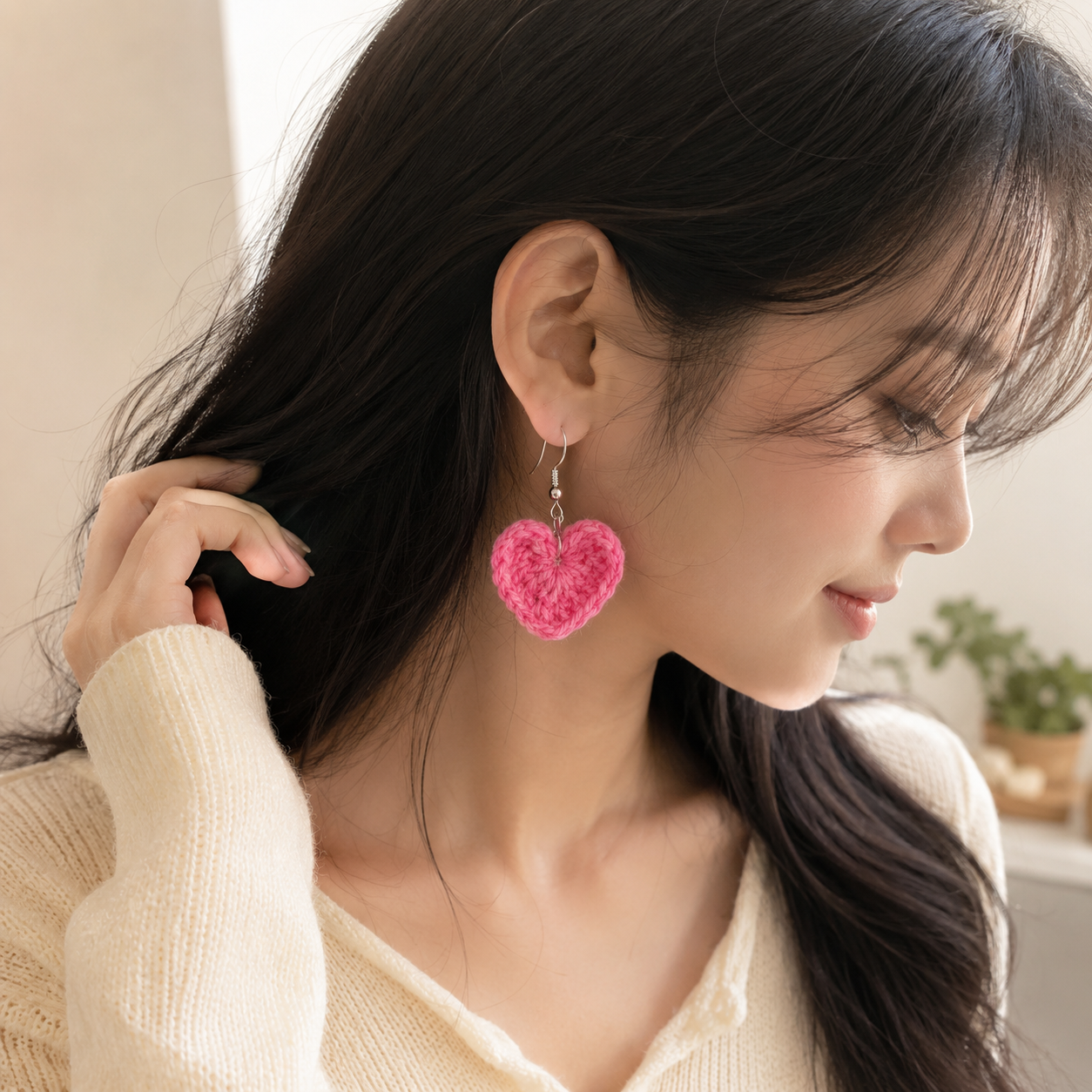 A woman wearing a bright pink crocheted heart-shaped dangle earring, shown in profile with her hand near her hair. She is wearing a cream-colored knit sweater in a softly lit indoor setting.