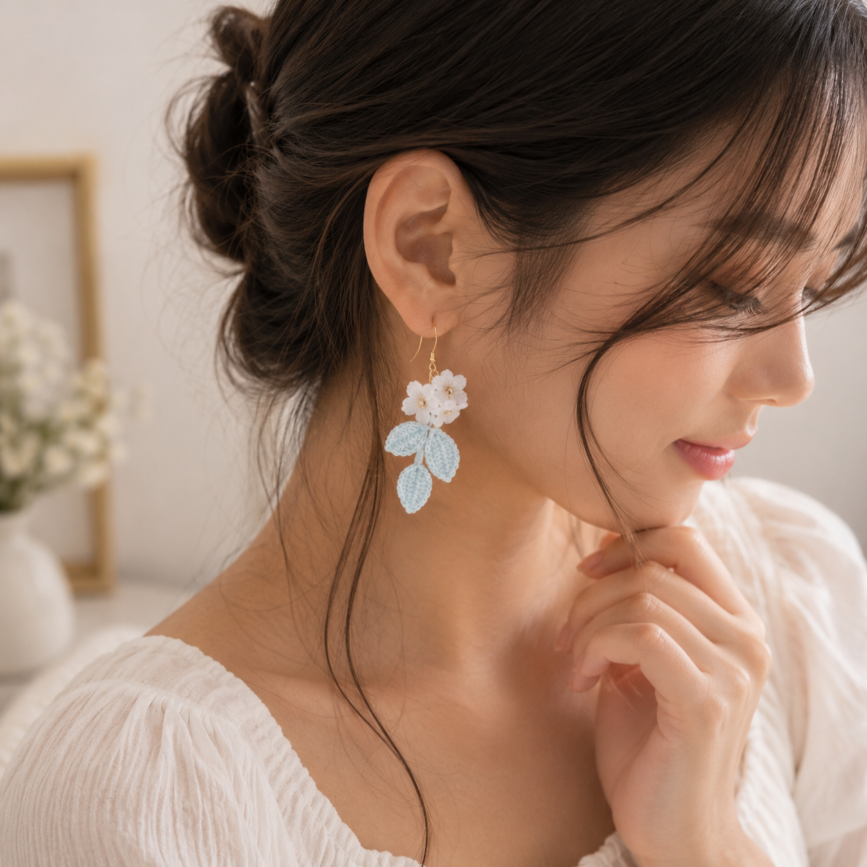 A woman wearing a delicate gold hoop earring adorned with a white flower and light blue leaves, shown in a soft, bright indoor setting.