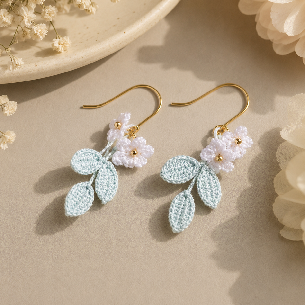 A pair of handmade crochet dangle earrings with light blue leaves, white flowers, and gold hooks, displayed on a neutral beige surface with dried flowers.