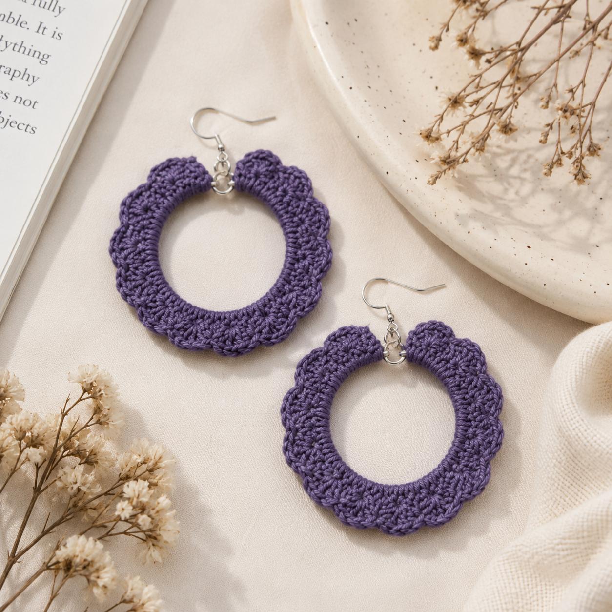 A pair of handmade purple crocheted hoop earrings with silver fishhook ear wires, displayed on a light beige textured surface with dried flowers and a ceramic plate.
