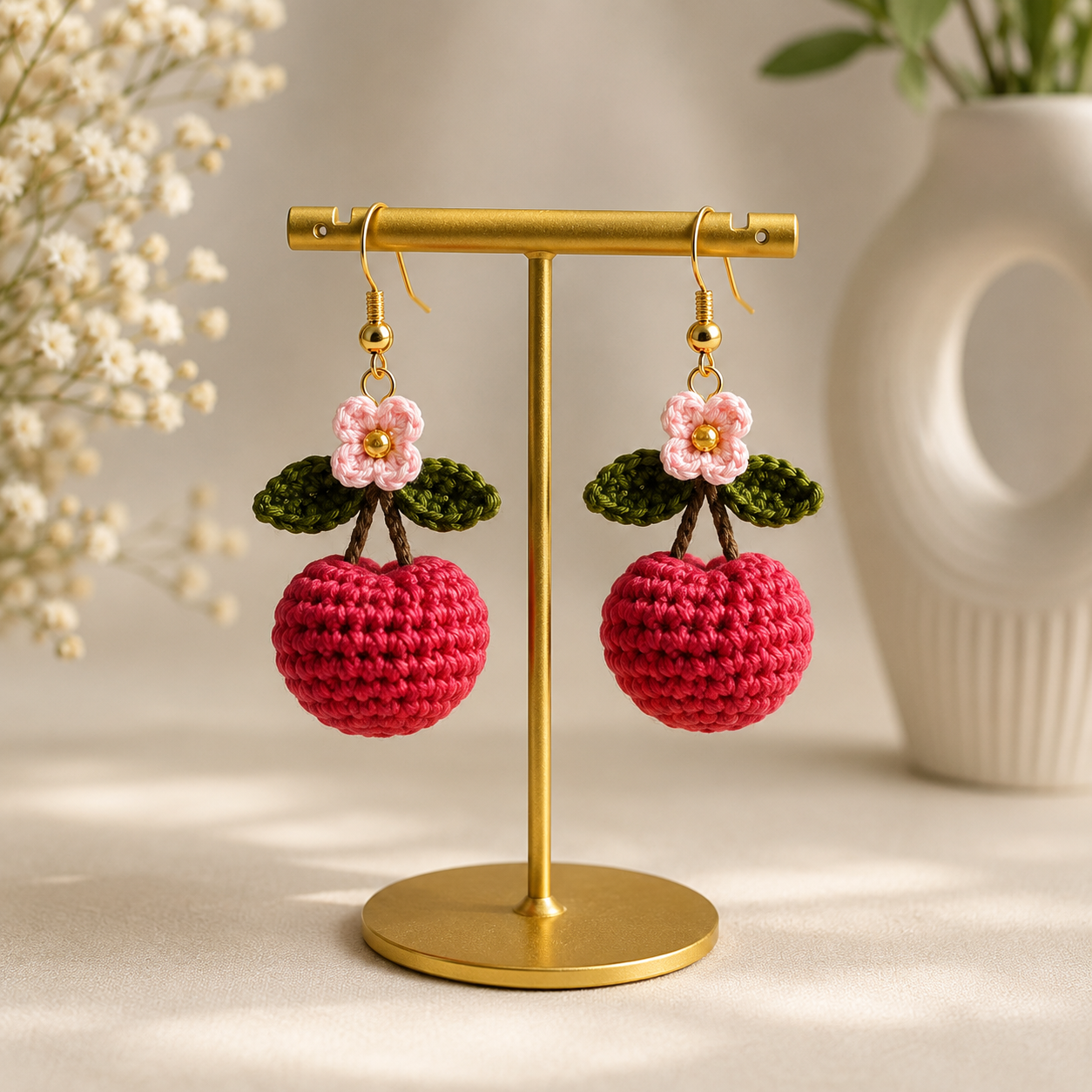 A pair of handmade crochet earrings shaped like red cherries with pink flowers and green leaves, hanging from a gold T-stand against a softly blurred beige background with dried flowers and a white vase.
