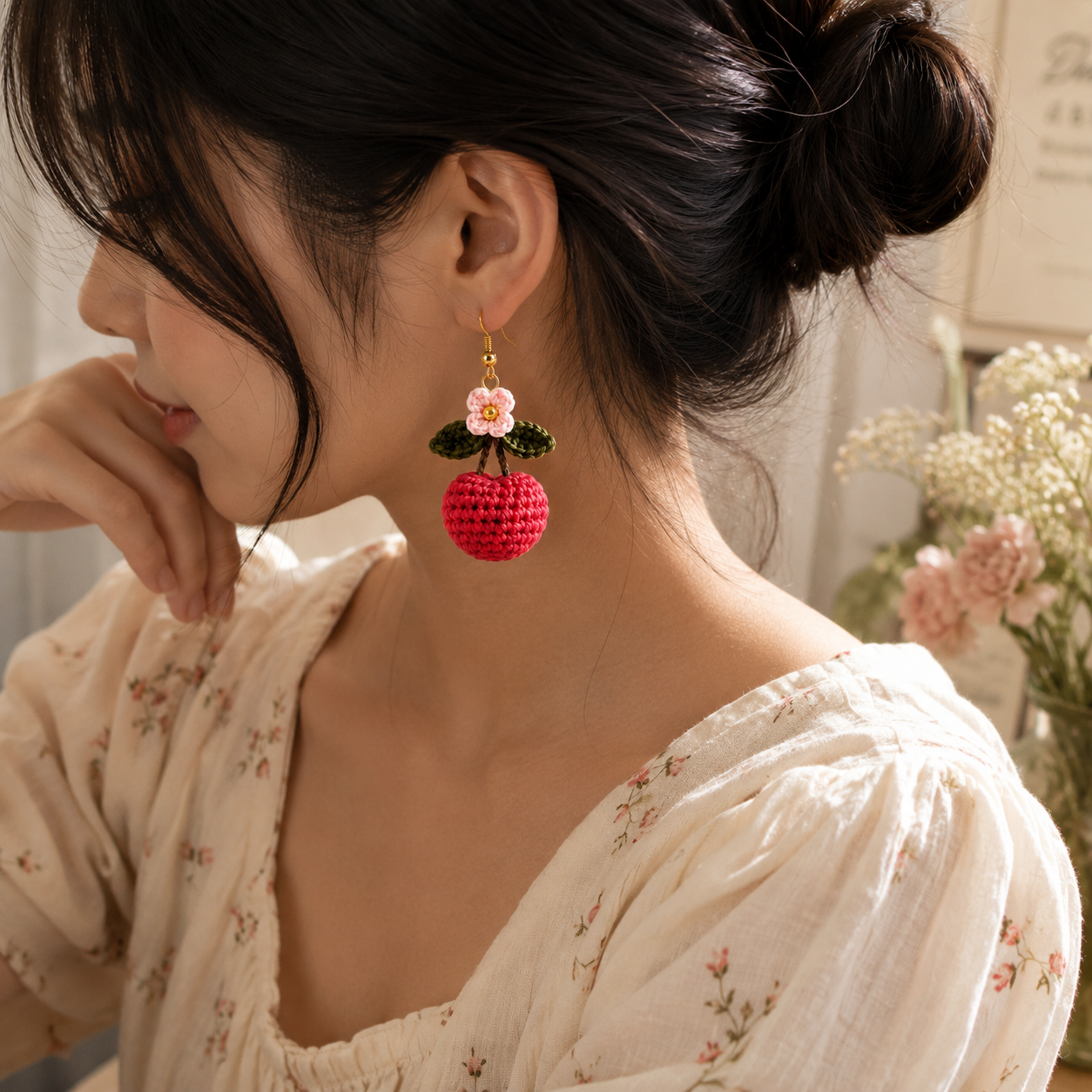 A woman wearing a pair of handmade crochet cherry drop earrings, shown in profile. The earrings feature a bright red crochet cherry body with green crochet leaves and a small white crochet flower with a gold-tone top hook.