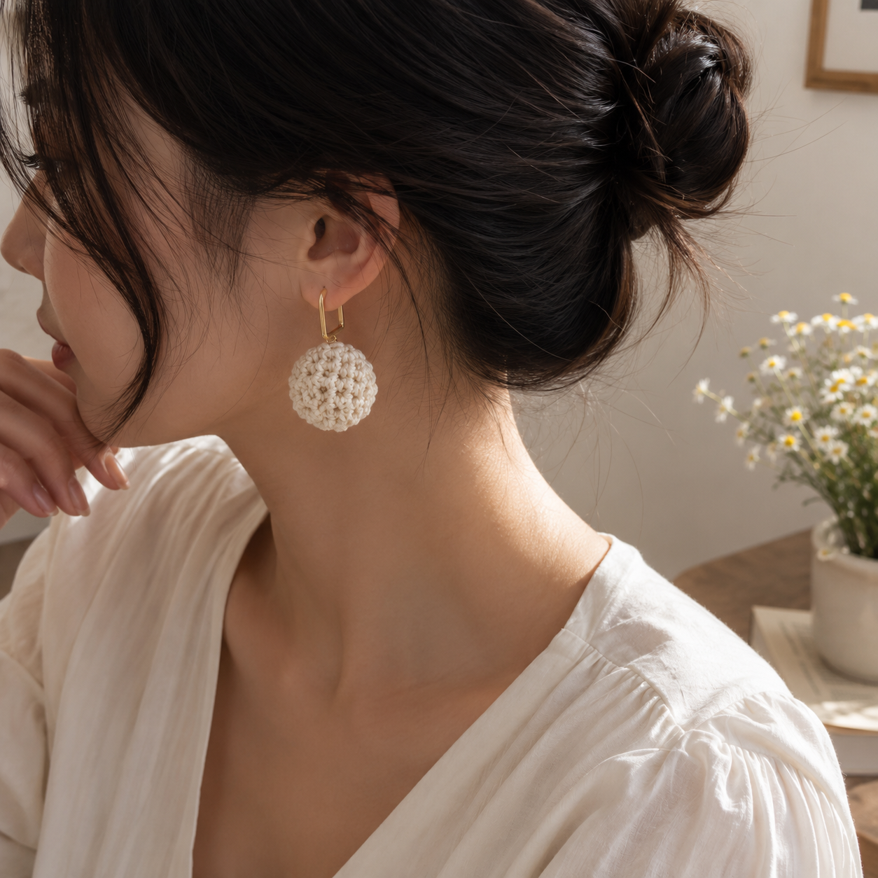 A woman wearing a white crocheted spherical drop earring with a gold hoop, shown in a close-up side profile in a softly lit indoor setting.