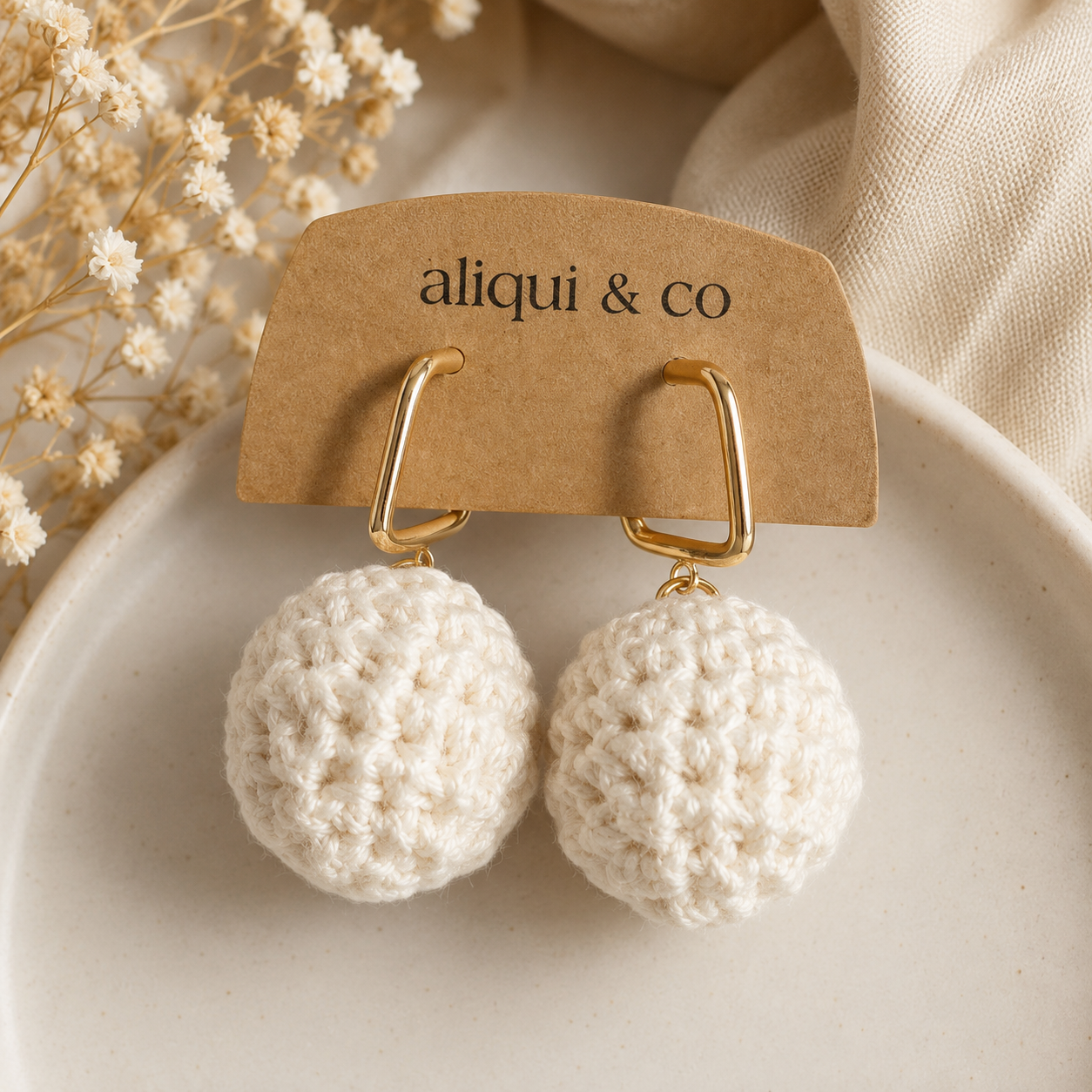A pair of cream crocheted pom-pom drop earrings with gold huggie hoops, displayed on a brown kraft paper card printed with the brand name 'aliqui & co'. The card is set on a light beige ceramic plate with dried flowers and a neutral fabric background.