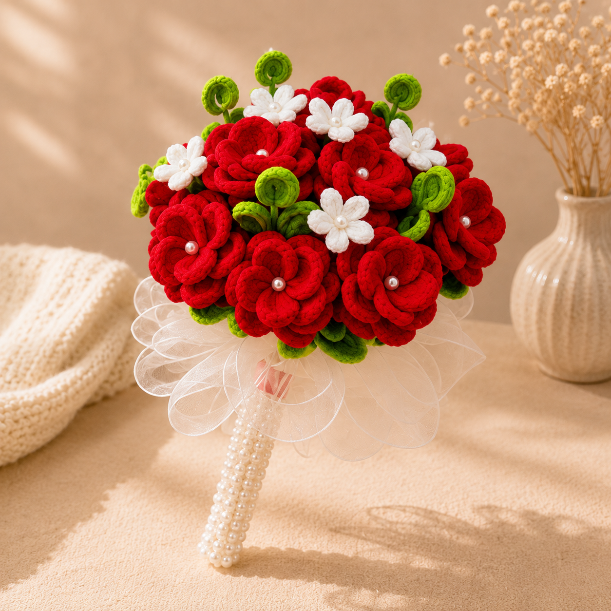 A handmade felt bouquet with red and white flowers, green leaves, and a pearl-wrapped handle, styled on a beige surface with a knitted blanket and dried flowers in the background.