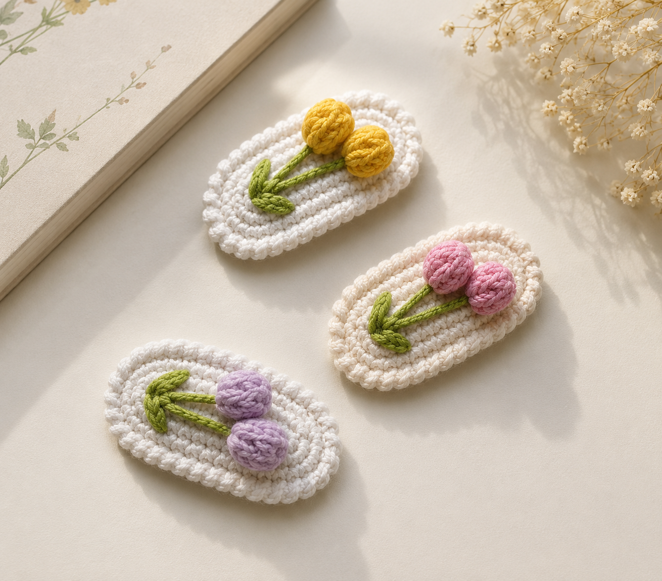 Three white crocheted hair clips with embroidered flowers, arranged on a light beige surface with dried flowers and a book corner in the background. The clips feature yellow, pink, and purple flowers with green stems.