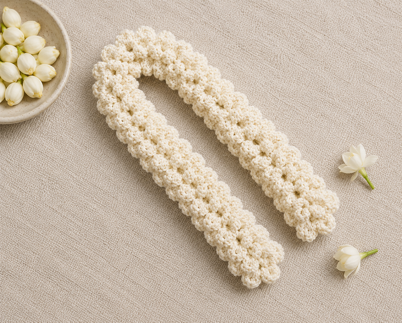 A cream-colored crocheted dishcloth with a textured floral pattern, laid flat on a beige linen surface with small white flowers and a bowl of flowers as props.