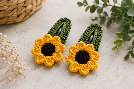 Two crocheted hair clips designed like sunflowers, featuring bright yellow petals, black centers, and dark green textured backs, displayed on a light textured surface with decorative greenery and a woven basket.
