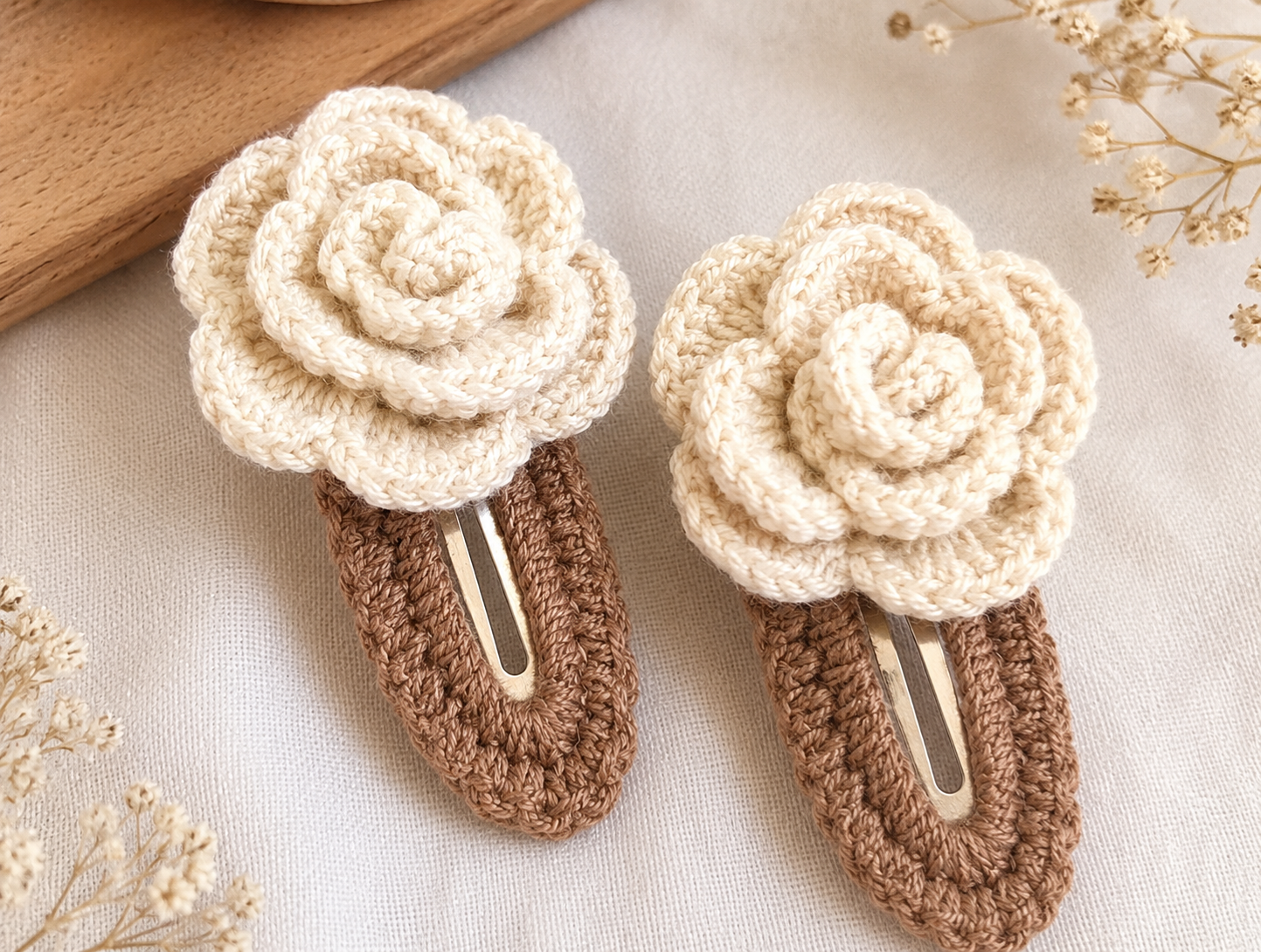 Two handmade crochet hair clips styled as cream-colored roses with brown textured bases, displayed on a light fabric background with dried flowers.