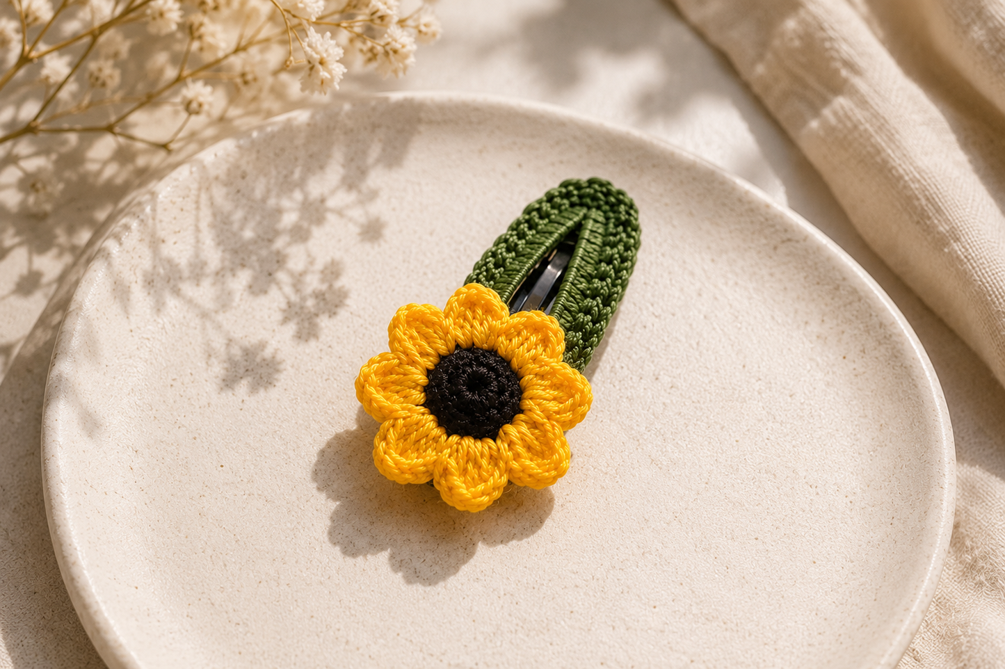 A crocheted sunflower hair clip with a dark green stem and a bright yellow flower with a black center, displayed on a textured beige ceramic plate.