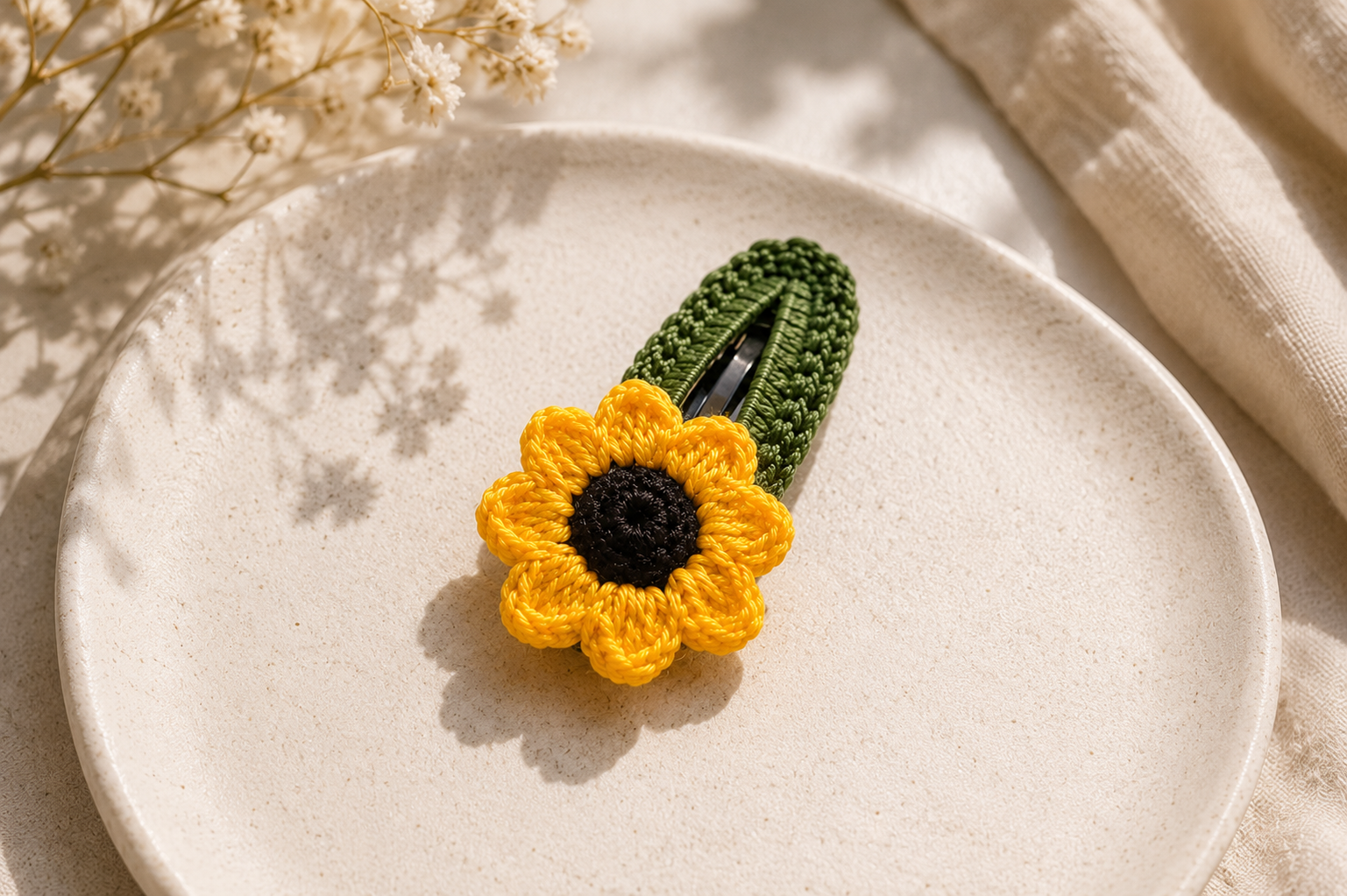 A crocheted sunflower hair clip with a dark green stem and a bright yellow flower with a black center, displayed on a textured beige ceramic plate.